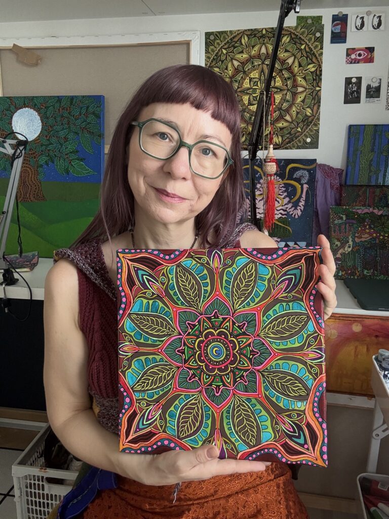Na zdjęciu autorka prezentuje w swoich rękach obraz mandala namalowana farbami akrylowymi z efektem świecenia pod lampą fluorescencyjną.